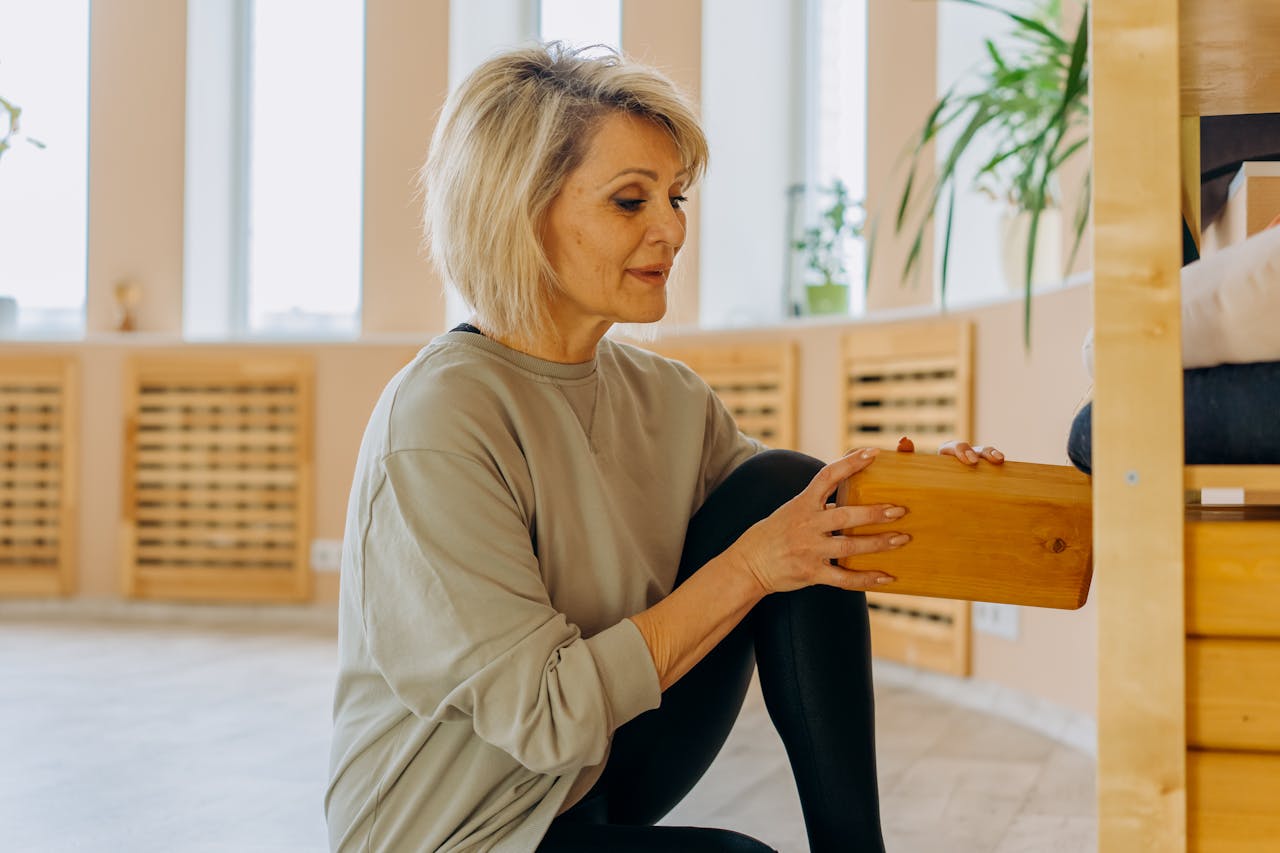 Mature woman using a yoga block indoors surrounded by natural light and plants.
